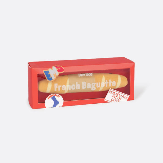 A red rectangular box containing a French baguette sock, photographed on a clean light gray background. The box has a clear front window and printed graphics including the words 'French Baguette' and small decorative badges.