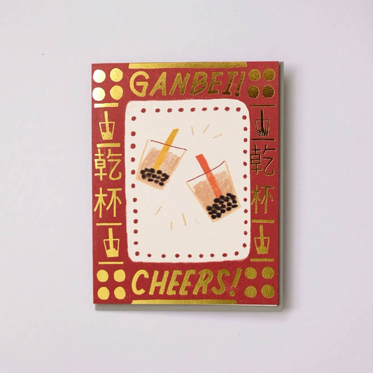 greeting card with gold foil text and symbols around the border. Center has illustrations of two boba cups. Text up top reads "Ganbei!" which is a form of "cheers" or "bottoms up" in Chinese. Bottom text reads "Cheers!" 