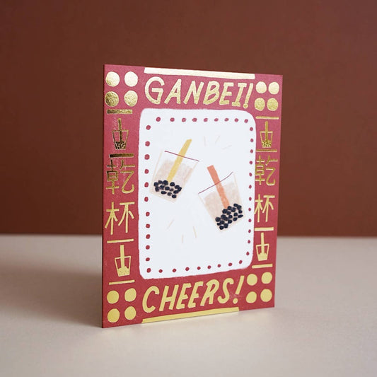 greeting card with gold foil text and symbols around the border. Center has illustrations of two boba cups. Text up top reads "Ganbei!" which is a form of "cheers" or "bottoms up" in Chinese. Bottom text reads "Cheers!" 