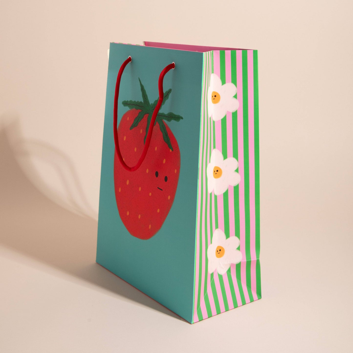 gift bag with a single giant strawberry on a turquoise background and side panel are white and yellow flowers on a pink and green vertical stripe background  