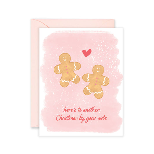 Holiday greeting card with two gingerbread men on it and a heart in between them. Text at the bottom reads "Here's to another Christmas by your side" 