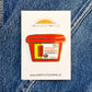 Sticker of a red container with yellow accents on a white background, placed on blue denim fabric.