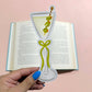 Hand holding a decorative bookmark shaped like a martini glass with olives on a pink background.