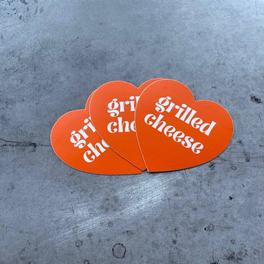 Orange heart shaped sticker with text that reads: Grilled Cheese. Three stickers.