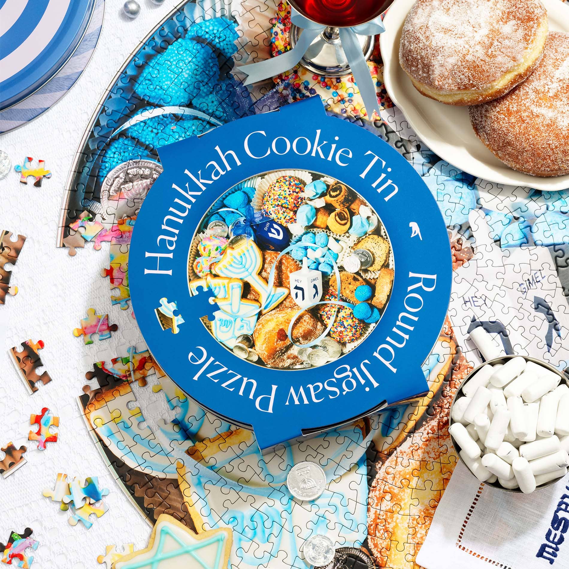 Round cookie tin that is blue and white stripe. Outer cover says Hanukkah Cookie Tin Jigsaw Puzzle with a photo of traditional cookies and dreidel.  Table is staged with jelly donuts and wine.