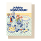 A rectangular greeting card with a illustrated Hanukkah table scene and the words “HAPPY HANUKKAH” at the top, shown with a kraft envelope behind it on a white background.