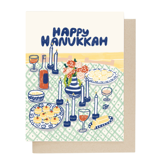 A rectangular greeting card with a illustrated Hanukkah table scene and the words “HAPPY HANUKKAH” at the top, shown with a kraft envelope behind it on a white background.