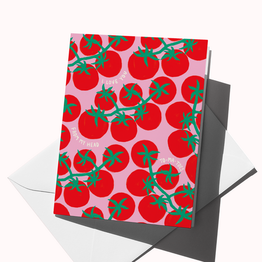 A square greeting card with a repeating tomato vine pattern in red and green on a light pink background, shown standing upright with a white envelope behind it on a clean white studio background.