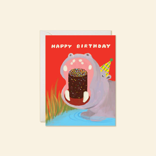 A square birthday greeting card with a red background and an illustration of a hippopotamus holding a chocolate cake with sprinkles. The text 'HAPPY BIRTHDAY' appears at the top, and a white envelope is partially visible behind the card.