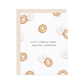 A white greeting card with a light beige envelope behind it, printed with scattered bagel illustrations and the phrase “LET’S SPREAD SOME HOLIDAY SCHMEAR” in gray text.