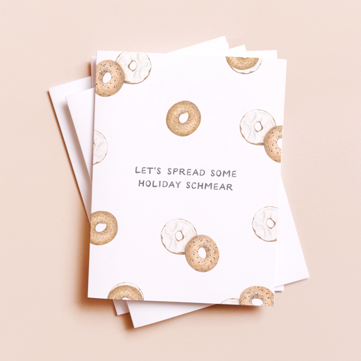 A stack of white greeting cards with a pattern of watercolor bagels and the text 'LET'S SPREAD SOME HOLIDAY SCHMEAR' on a light beige background.