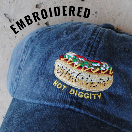 Close Up Photo: Faded blue baseball hat with embroidery of hot dog with poppy seed bun and chicago-style toppings. Yellow embroidered text underneath hot dog says "Hot Diggity."