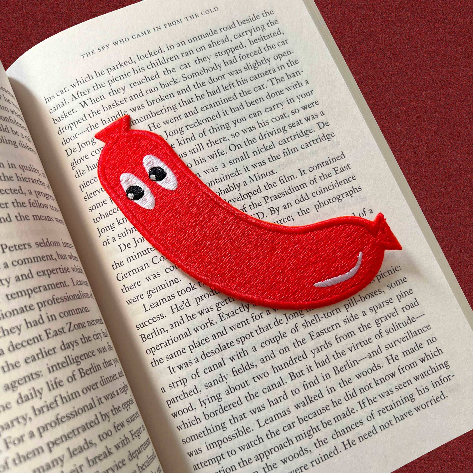 Embroidered fabric patch shaped like a frankfurter or hotdog (without the bun). Red with cartoonish eyes looking off to the side.