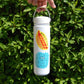 A hand holds a white insulated water bottle with a carrying loop against a background of green leaves. The bottle features a cartoon hot dog graphic with 'hottie' text and a blue bubbly character below it.
