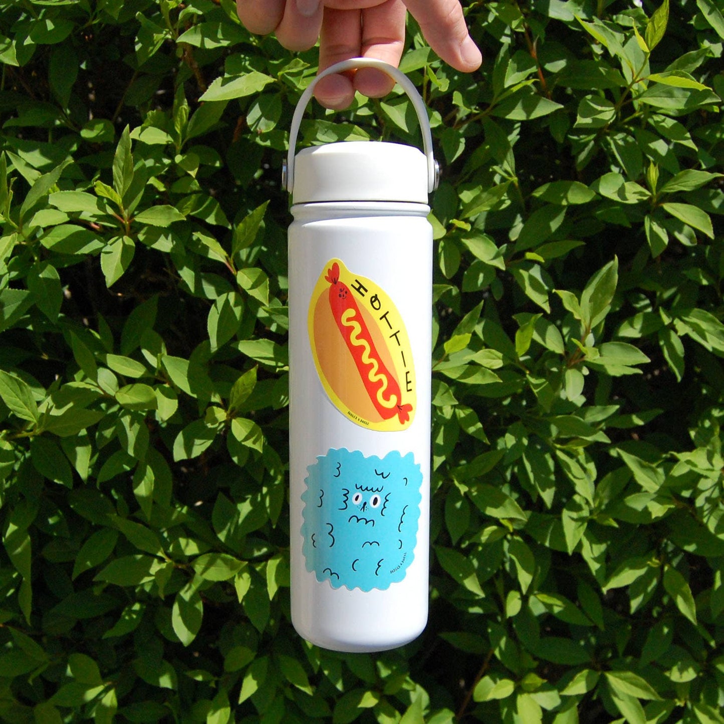 A hand holds a white insulated water bottle with a carrying loop against a background of green leaves. The bottle features a cartoon hot dog graphic with 'hottie' text and a blue bubbly character below it.