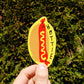 A hand holds a die-cut sticker shaped like a hot dog in a bun, with the word 'HOTTIE' printed vertically. The sticker has a yellow bun, a red hot dog with yellow mustard, and black text.