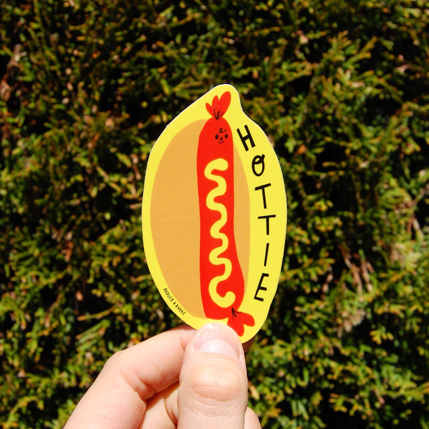 A hand holds a die-cut sticker shaped like a hot dog in a bun, with the word 'HOTTIE' printed vertically. The sticker has a yellow bun, a red hot dog with yellow mustard, and black text.