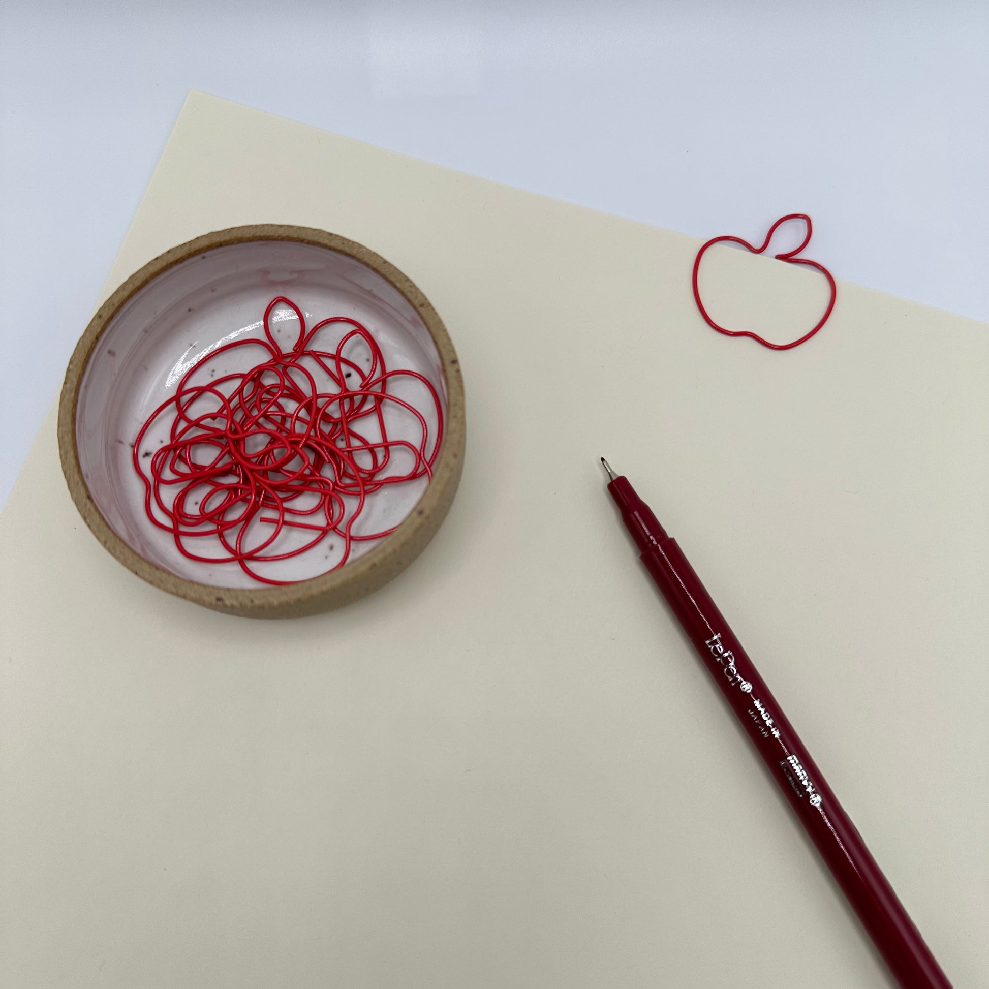 Staged Paperclips with paper, ramekin holding red apple paperclips, and burgundy Le Pen.