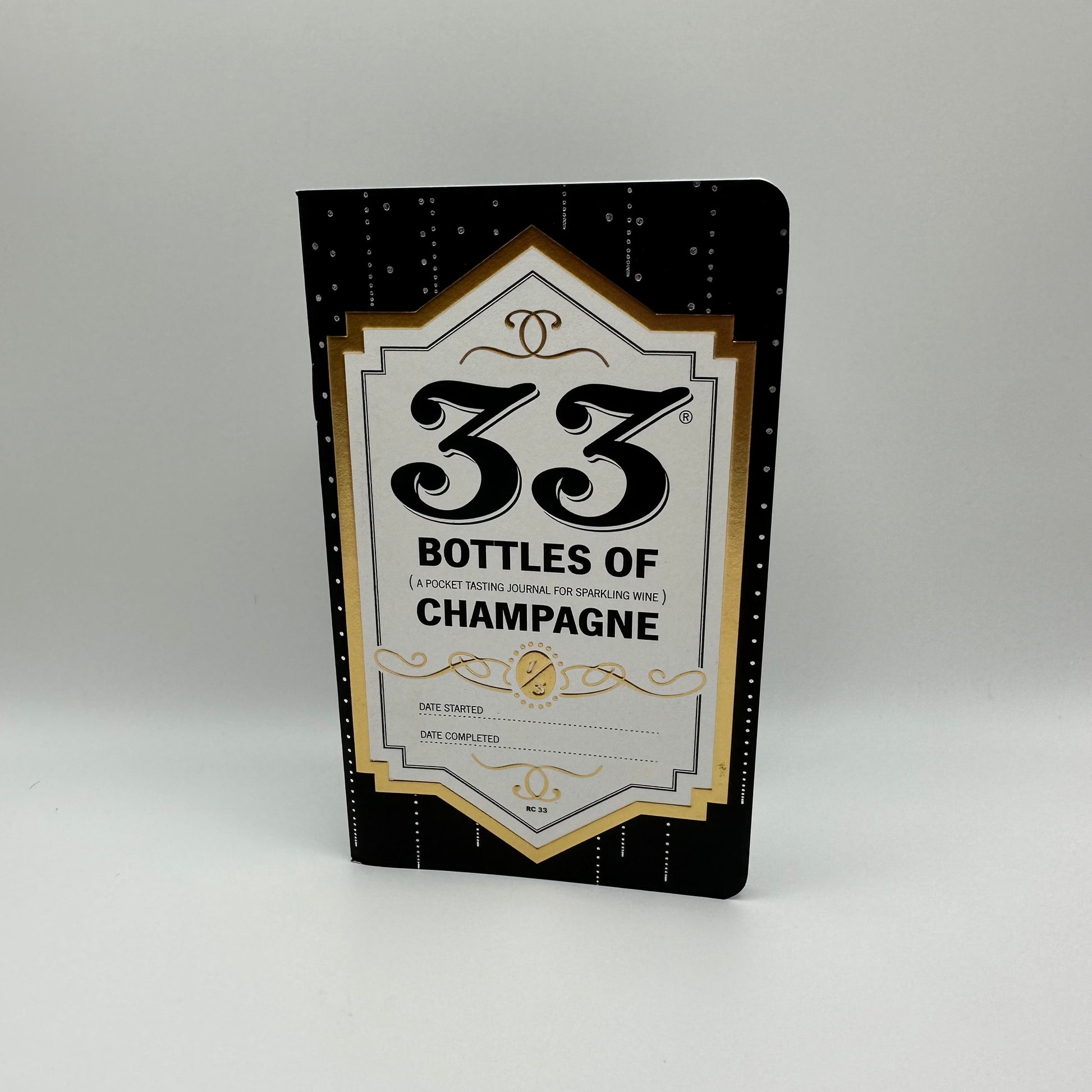 Photo depicting front cover of journal on white background. Journal is recycled paper dyed black with gold trim and black text title "33 Bottles of Champagne - A Pocket Tasting Journal for sparkling wine." Also small text on the bottom for user to notate "Date Started" and "Date Completed.".