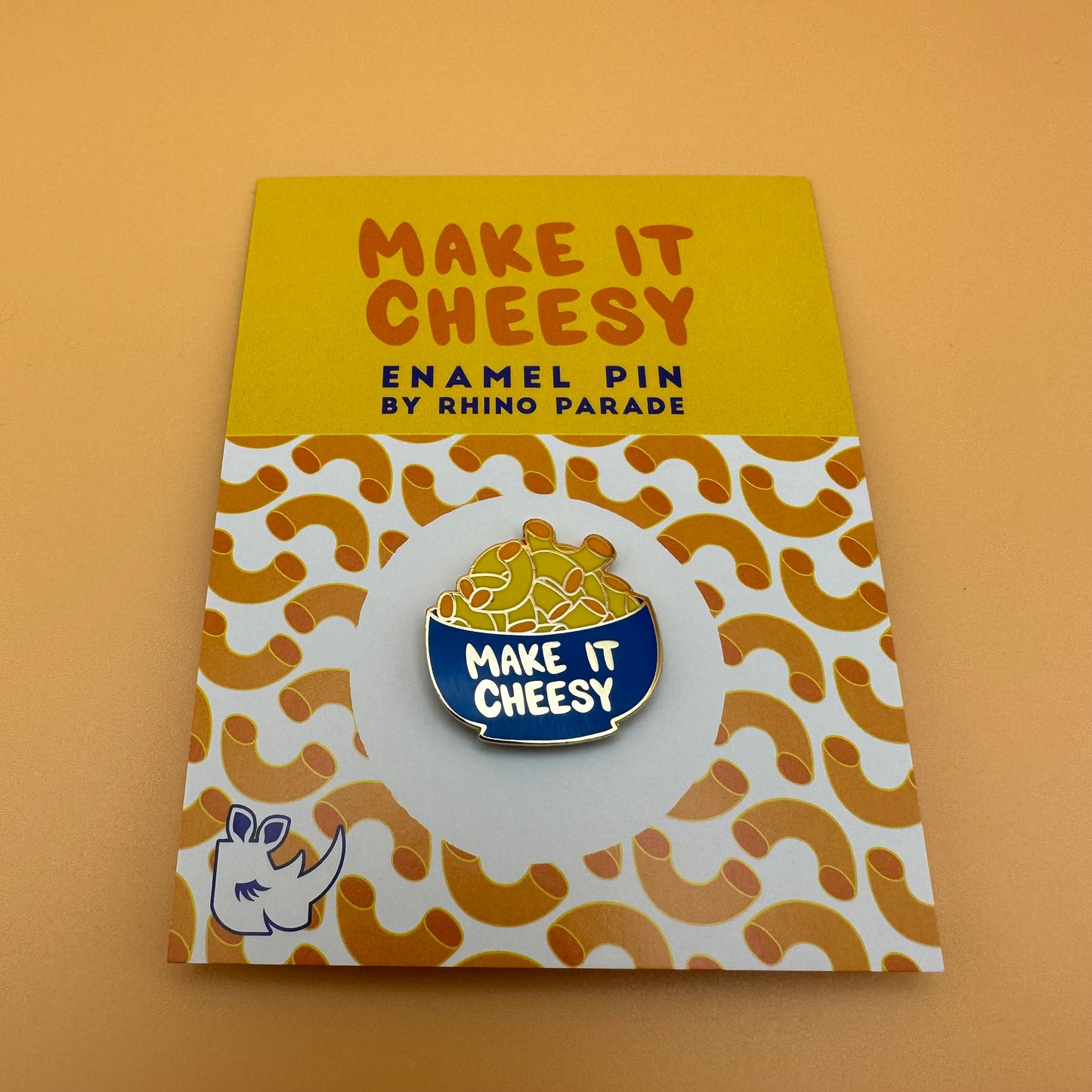 Bowl of mac and cheese enamel pin that reads "make it cheesy." On a card backing that has a patterned image of macaroni moodles.