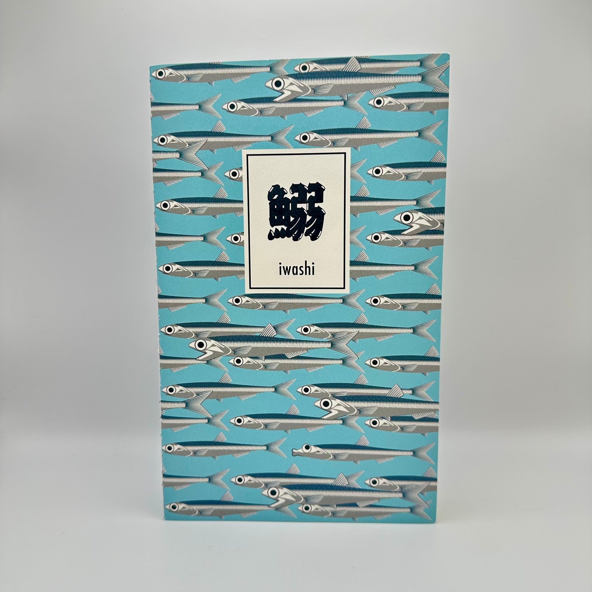 Notebook with blue background with silver sardines swimming. Text box in Japanese that reads "Iwashi"