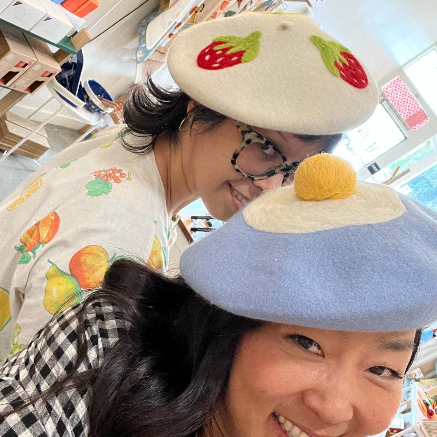 2 Ladies modeling a white beret with strawberries and a blue beret with an egg.