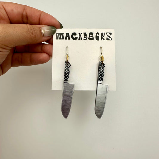 Hand holding a card with 2 earrings that look like knives. The handles are black and white checker. The card says Mackbecks.