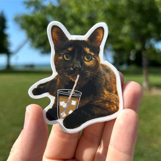 Sticker of a cat holding a drink with a straw, held in a hand outdoors.