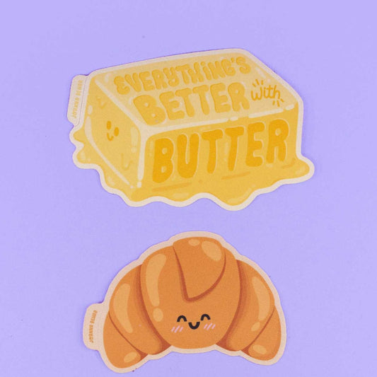 Two stickers on a purple backdrop. One of butter that says Everything's Better with Butter, and the second one that is of a croissant smiling.