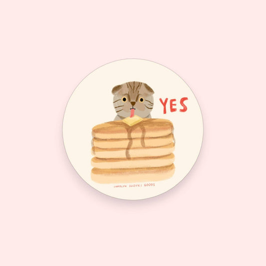 round sticker with illustrated cat licking pad of butter on a stack of pancakes and text just reads "yes" 