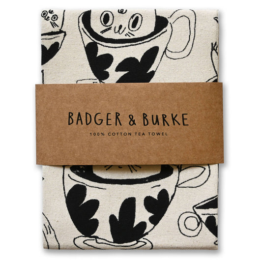 A folded cotton tea towel with a black and white coffee cup pattern, wrapped in a brown paper band that reads 'BADGER & BURKE 100% COTTON TEA TOWEL'.