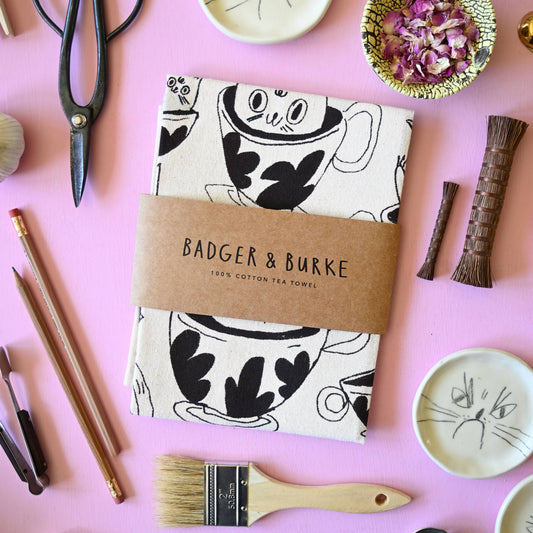 A folded cotton tea towel with a black and white cartoon teacup pattern, wrapped in a brown paper band reading 'BADGER & BURKE'. The towel is styled in a flat-lay on a pink background with tea-related props like scissors, a whisk, and teacups.