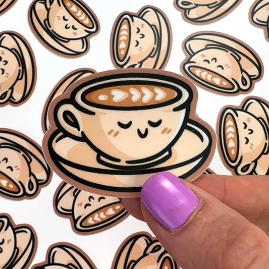 A hand with purple nail polish holds a die-cut sticker featuring a cute cartoon coffee cup with latte art and a smiling face, with more matching stickers in the background.