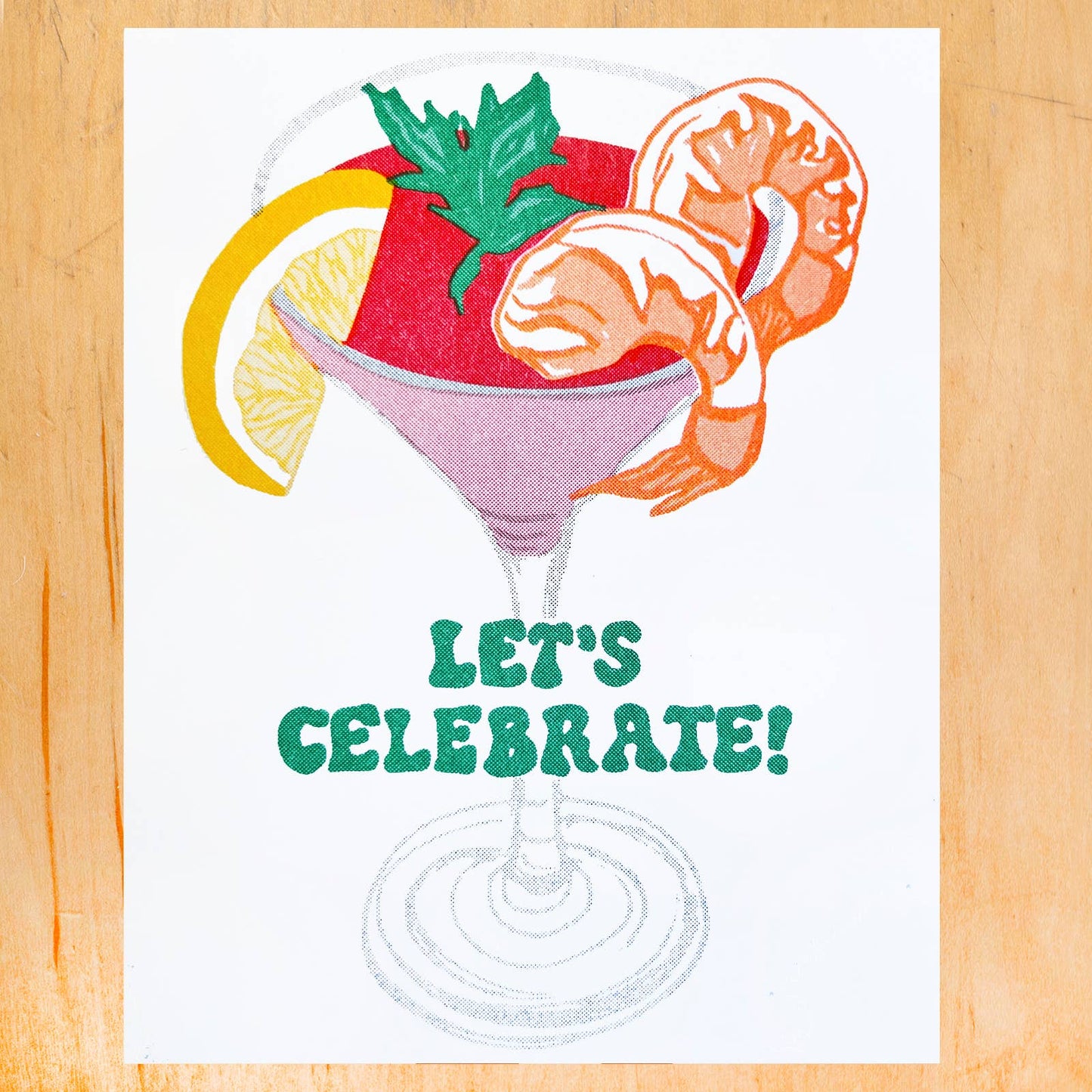 A square white art print or card featuring a colorful cocktail illustration with shrimp, a lemon slice, green garnish, and the text 'LET'S CELEBRATE!' in green, displayed on a light wood background.