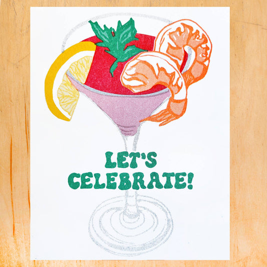 A square white art print or card featuring a colorful cocktail illustration with shrimp, a lemon slice, green garnish, and the text 'LET'S CELEBRATE!' in green, displayed on a light wood background.