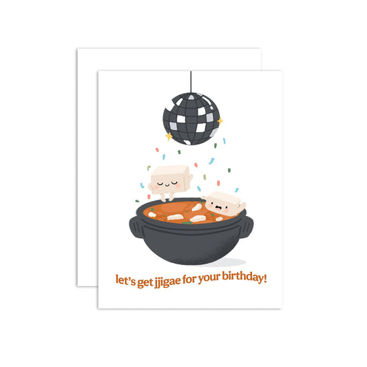 A greeting card with a white background featuring a cartoon illustration of two pieces of tofu in an bowl of korean soup, with a disco ball above and the text 'let's get jjigae for your birthday!' in orange. A white envelope is partially visible behind the front card.