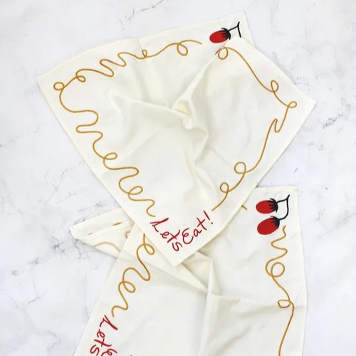 Two off-white napkins embroidered with a squiggly spaghetti noodle border, two red tomatoes, and "Let's Eat!" in red text. Shown on a marble surface.