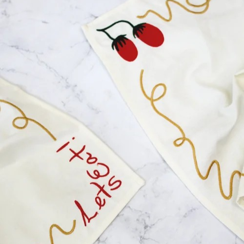 Two off-white napkins embroidered with a squiggly spaghetti noodle border, two red tomatoes, and "Let's Eat!" in red text. Shown on a marble surface. Detail shot of embroidery.
