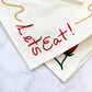 Two off-white napkins embroidered with a squiggly spaghetti noodle border, two red tomatoes, and "Let's Eat!" in red text. Shown on a marble surface. Detail shot of embroidery.