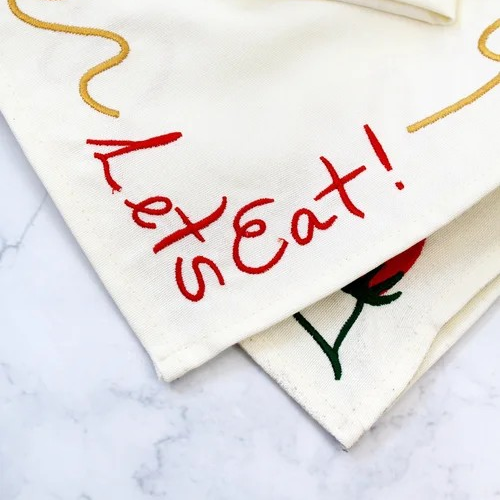 Two off-white napkins embroidered with a squiggly spaghetti noodle border, two red tomatoes, and "Let's Eat!" in red text. Shown on a marble surface. Detail shot of embroidery.