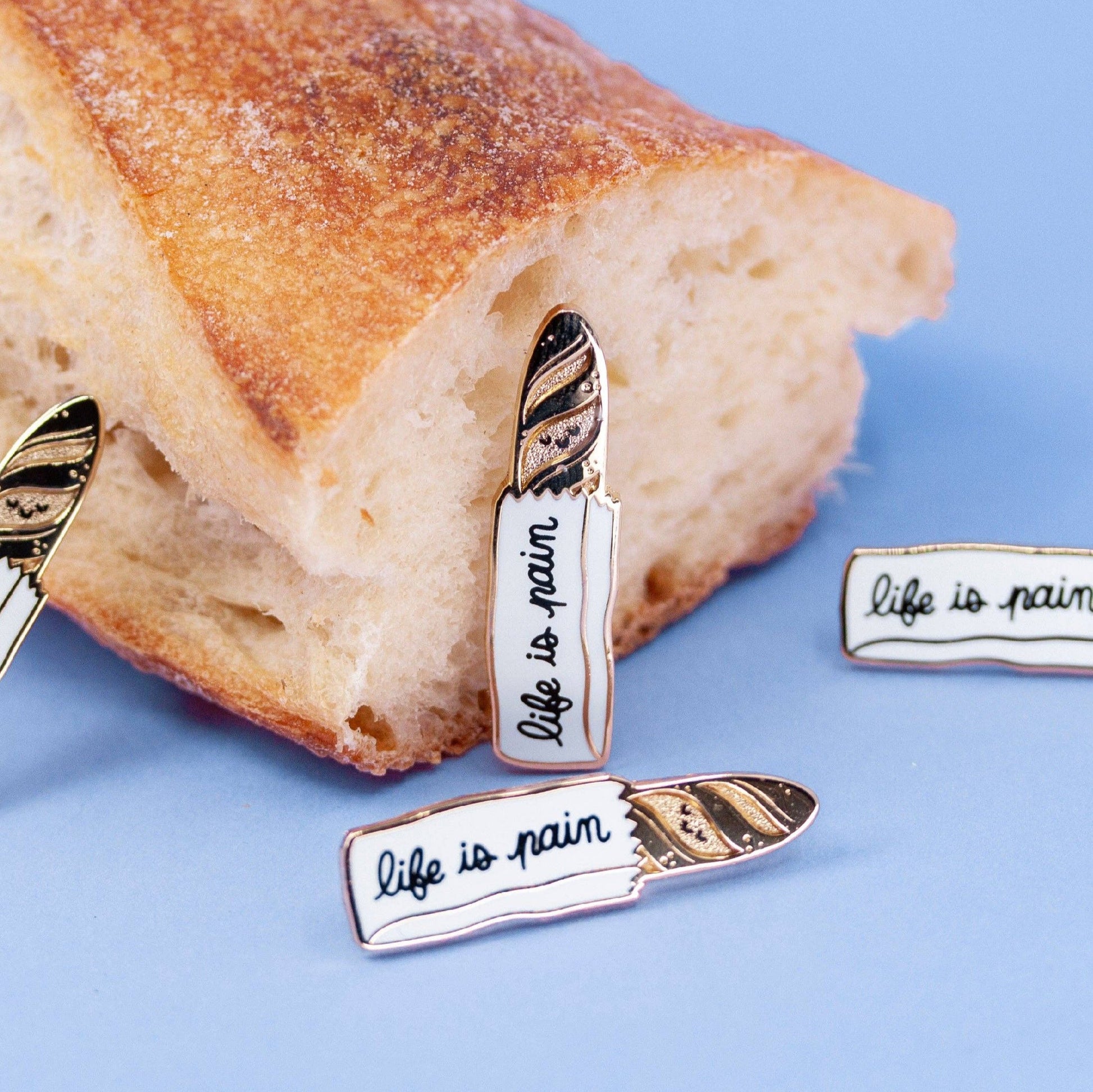 a lapel pin that is of a gold baguette sticking out of a bread bag. Text on the bag reads "life is a pain"