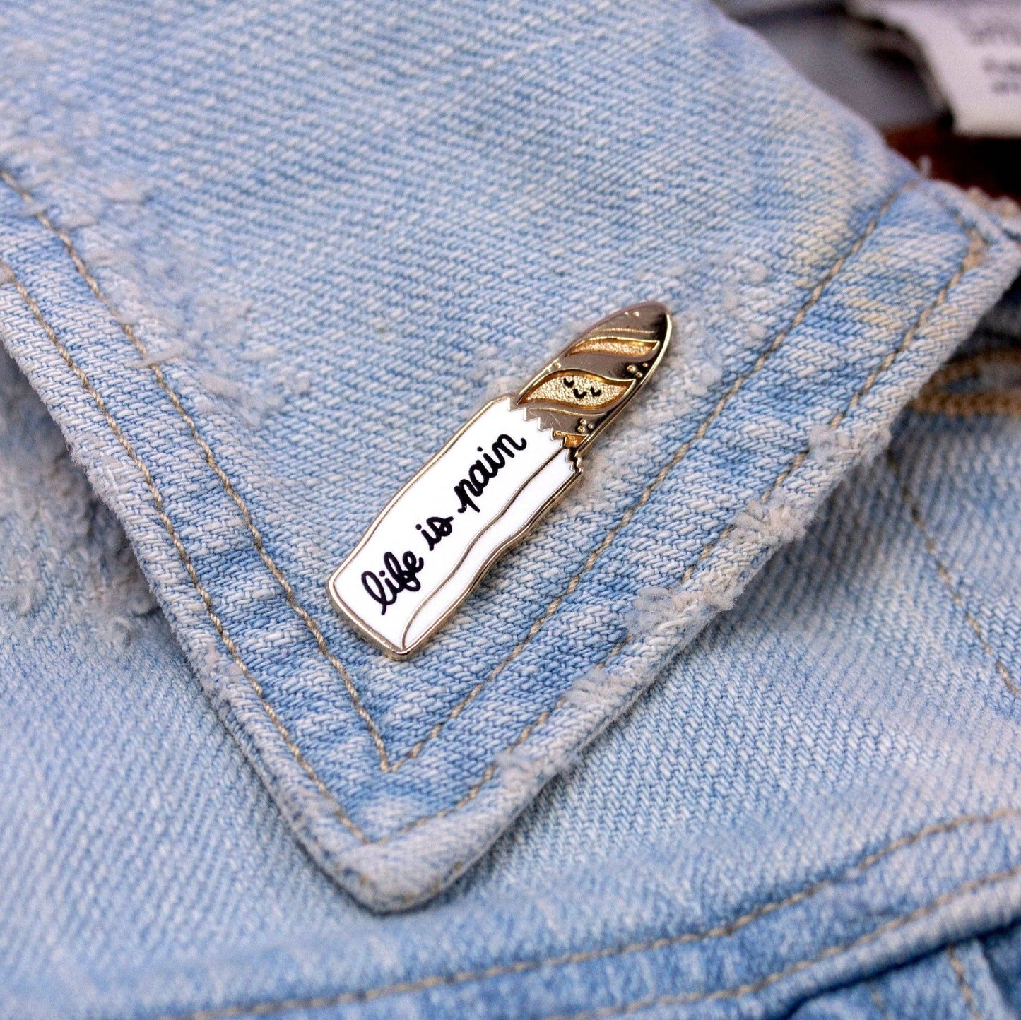 a lapel pin that is of a gold baguette sticking out of a bread bag. Text on the bag reads "life is a pain"