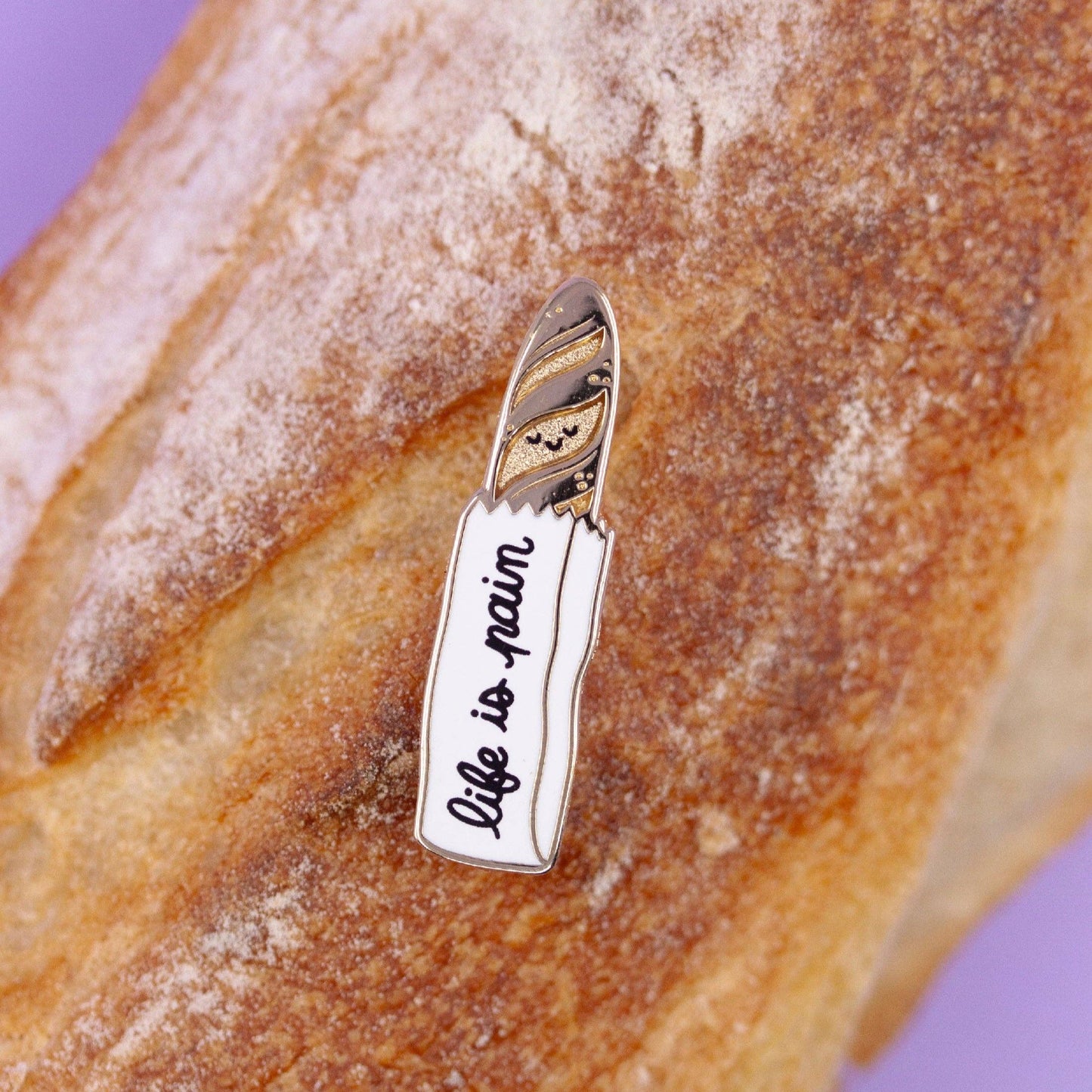 a lapel pin that is of a gold baguette sticking out of a bread bag. Text on the bag reads "life is a pain"