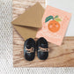 Pair of black baby shoes with tan laces on a wooden surface, next to a brown envelope and a greeting card with an orange design.