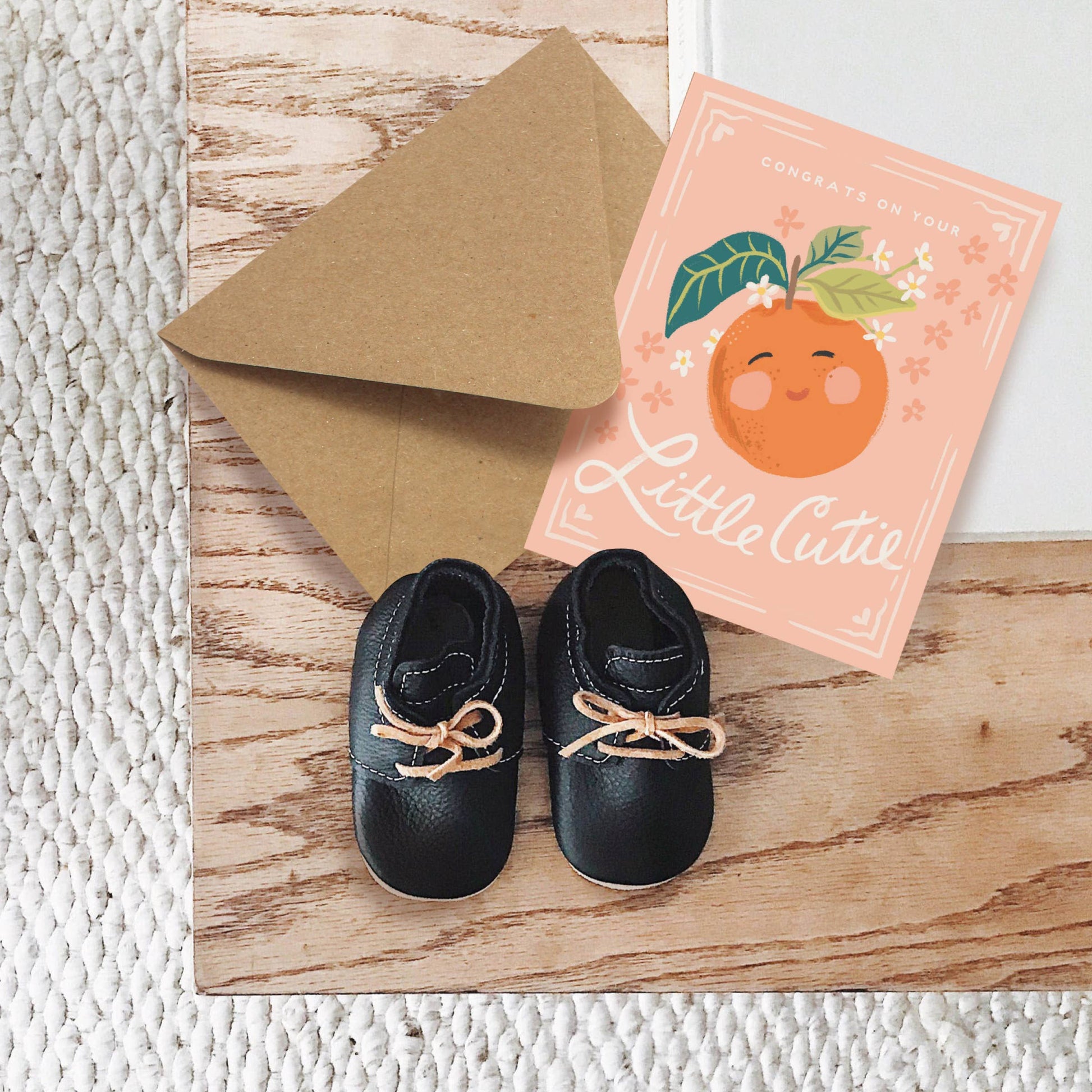 Pair of black baby shoes with tan laces on a wooden surface, next to a brown envelope and a greeting card with an orange design.