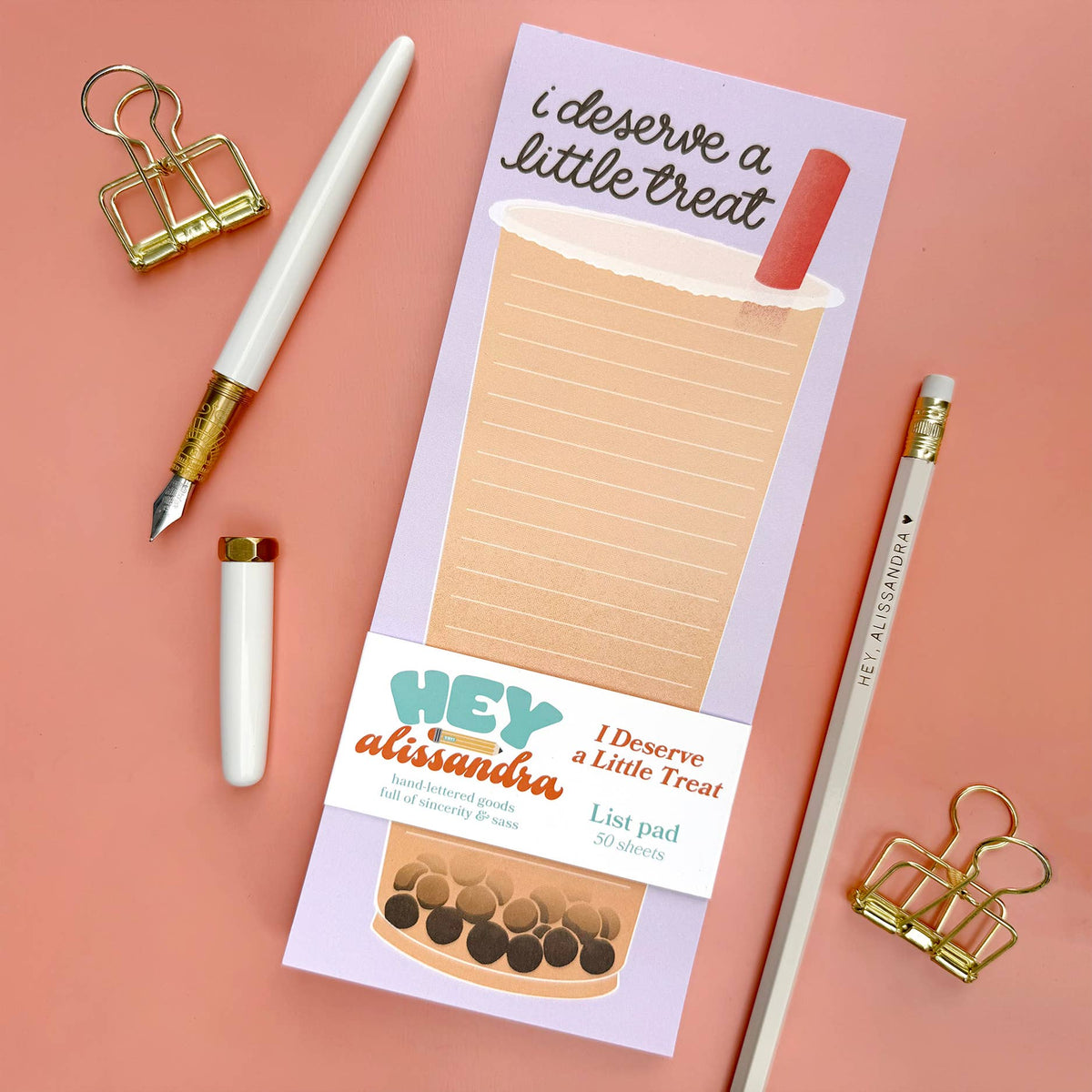 A pastel lavender list pad with a coffee cup illustration and the phrase 'i deserve a little treat' is shown on a pink background, styled with a white pen, gold binder clips, a pencil, and a small candle. The lower band reads 'HEY alissandra' and 'I Deserve a Little Treat.