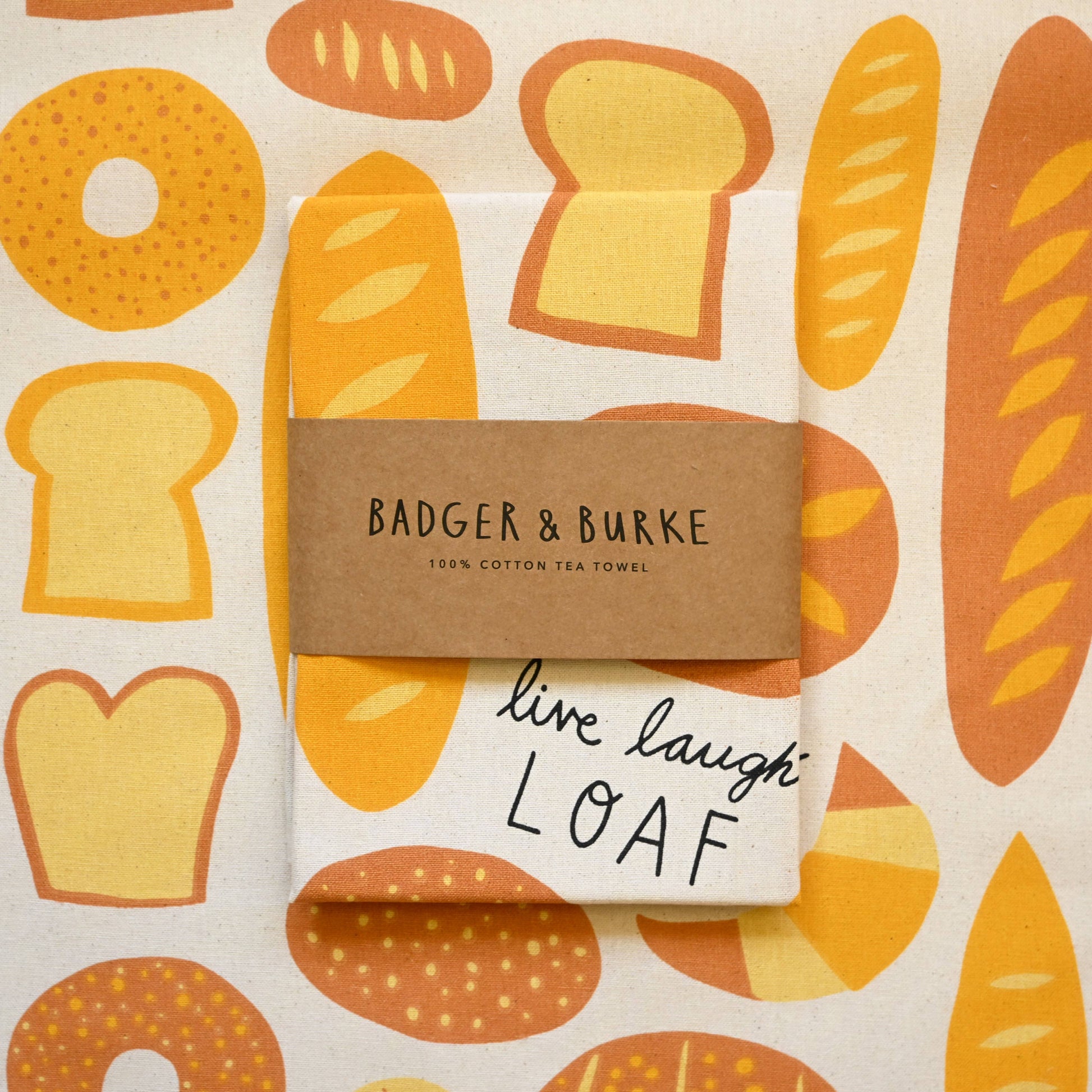 A folded cotton tea towel with a bread pattern, wrapped with a brown paper band reading 'BADGER & BURKE 100% COTTON TEA TOWEL' and handwritten 'live laugh LOAF' on the front.