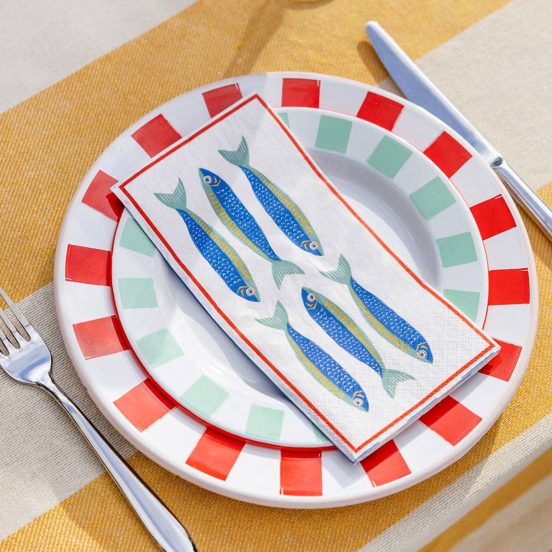 Decorative plate with striped rim, colorful fish design on a napkin, and cutlery on a yellow and beige checkered tablecloth.