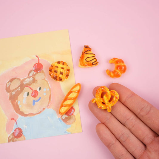 5 mini magnets that look like pastries. Hand is holding the pretzel magnet. Picture of a bear with cherries all over it for decor.