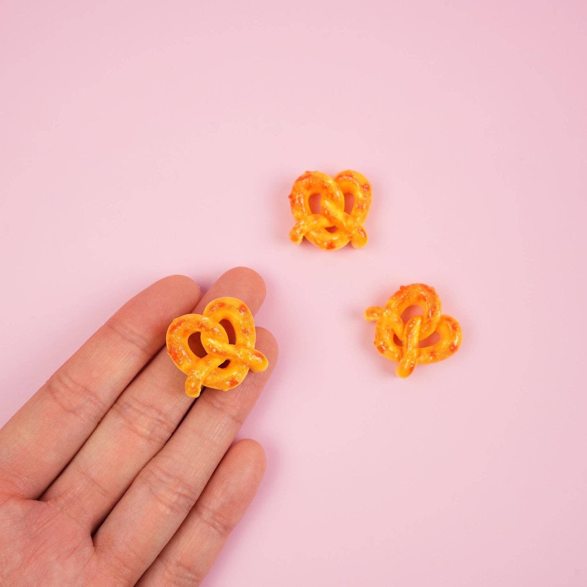 Hand holding pretzel magnet. 2 others are on a table with a pink backdrop.